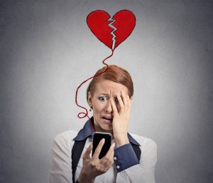 Sad woman with broken heart looking at her mobile phone isolated on grey wall background
